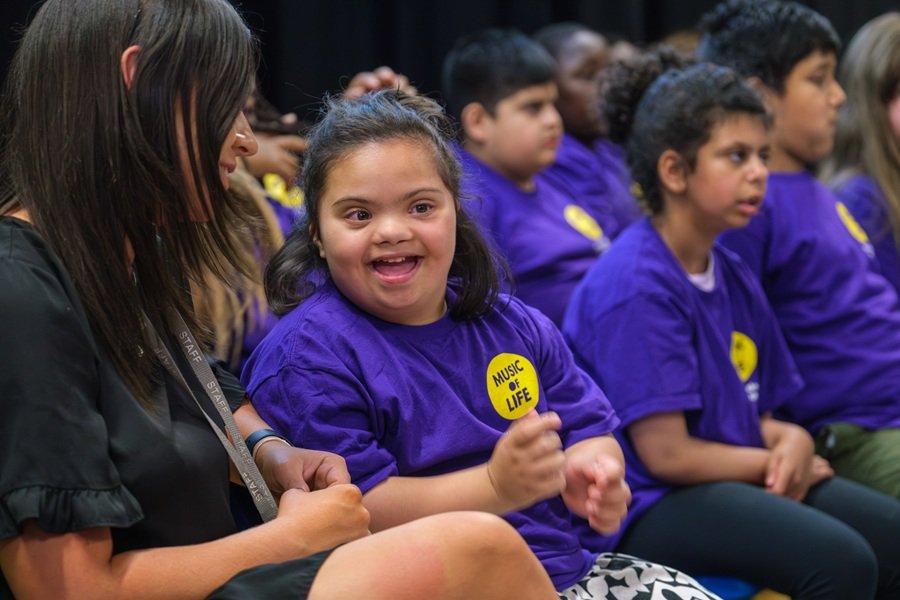 Young participants engage in a Music of Life session | Photo by Music of Life | Image provided by Music of Life A group of children and young people sit together during a Music of Life activity. In the foreground, one participant wearing a purple Music of Life T-shirt sits alongside an adult facilitator and gestures with their hands while engaging with the session. Other participants wearing matching purple T-shirts sit nearby, creating a group environment focused on participation, interaction and shared musical experiences.