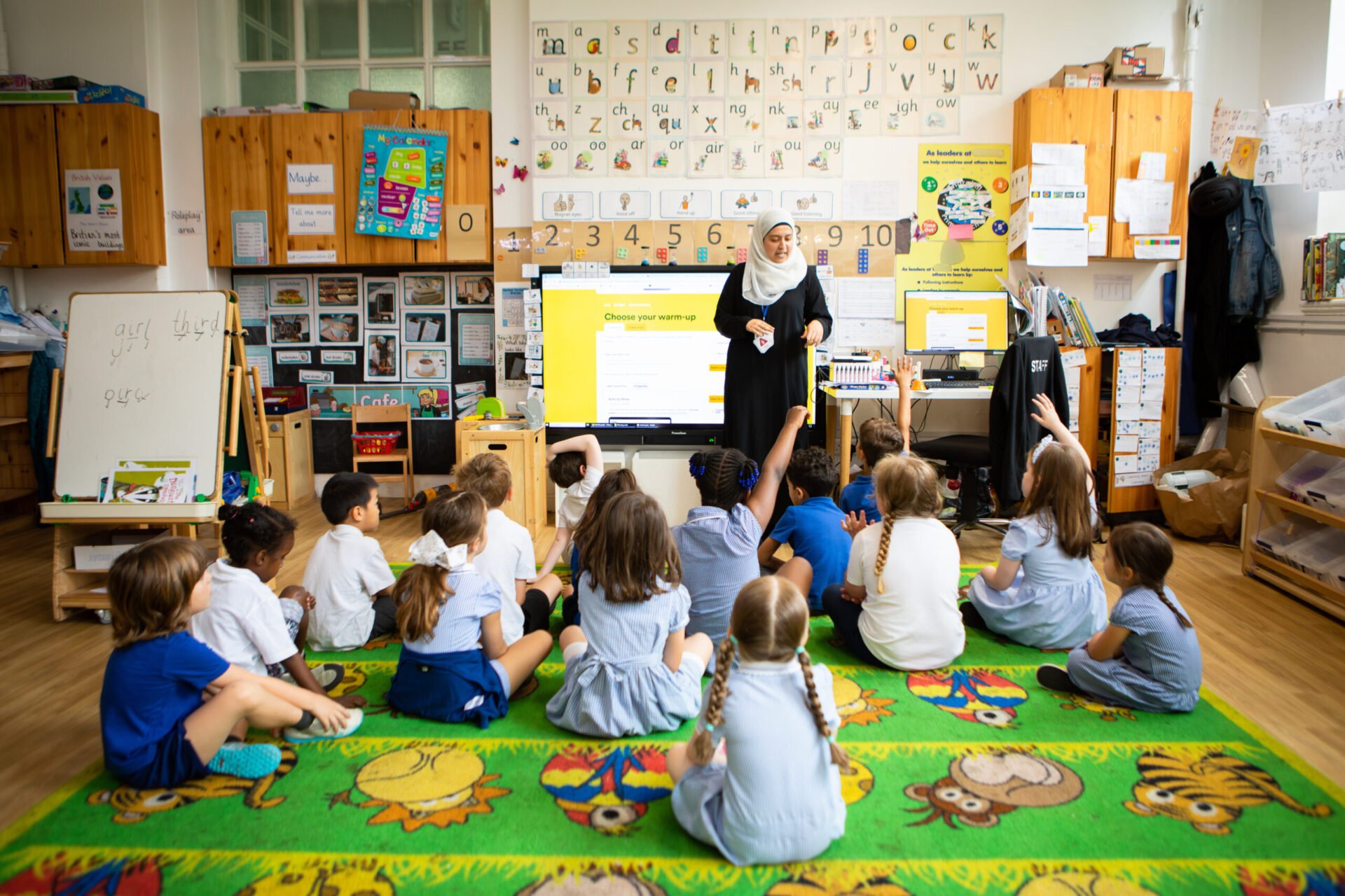Teacher invites pupils to choose a classroom warm-up | Photo by Stanton Media | Image provided by Aurora Orchestra A teacher stands beside a large classroom screen displaying the words “Choose your warm-up”. In front of her, pupils in school uniform sit in a semicircle on a brightly coloured carpet, with several raising their hands to contribute.