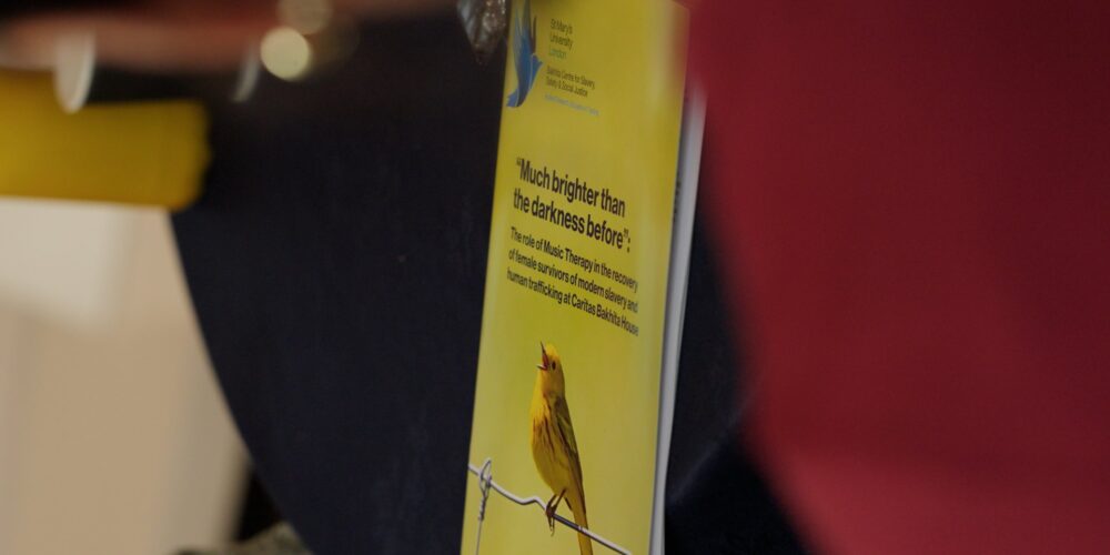 Research into music therapy at Caritas Bakhita House is launched | Photo by Diocese of Westminster Communications Office | Image provided by Caritas Bakhita House A copy of a research publication lies on a dark table during an event. The yellow cover features an illustration of a small bird perched on a wire and includes the title “Much brighter than the darkness before” with text describing music therapy in the recovery of female survivors of modern slavery and human trafficking at Caritas Bakhita House. The booklet is photographed in close-up, with people and objects blurred in the foreground and background.