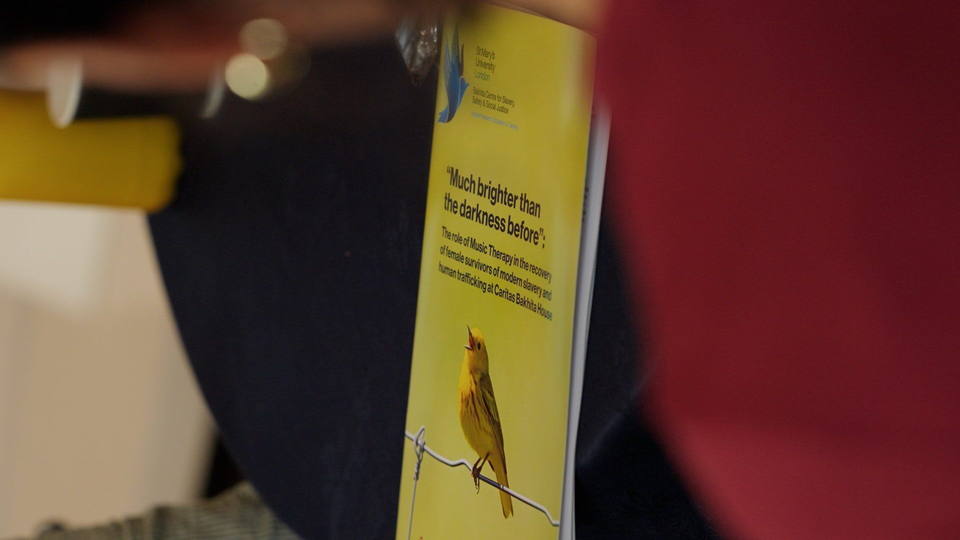 A copy of a research publication lies on a dark table during an event. The yellow cover features an illustration of a small bird perched on a wire and includes the title “Much brighter than the darkness before” with text describing music therapy in the recovery of female survivors of modern slavery and human trafficking at Caritas Bakhita House. The booklet is photographed in close-up, with people and objects blurred in the foreground and background.